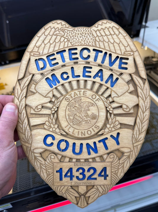 A hand holds a large, laser-engraved wooden replica badge of a Detective McLean, Illinois County. The badge is made of light-colored wood with blue painted lettering and numbers, and features an eagle and leaf design.