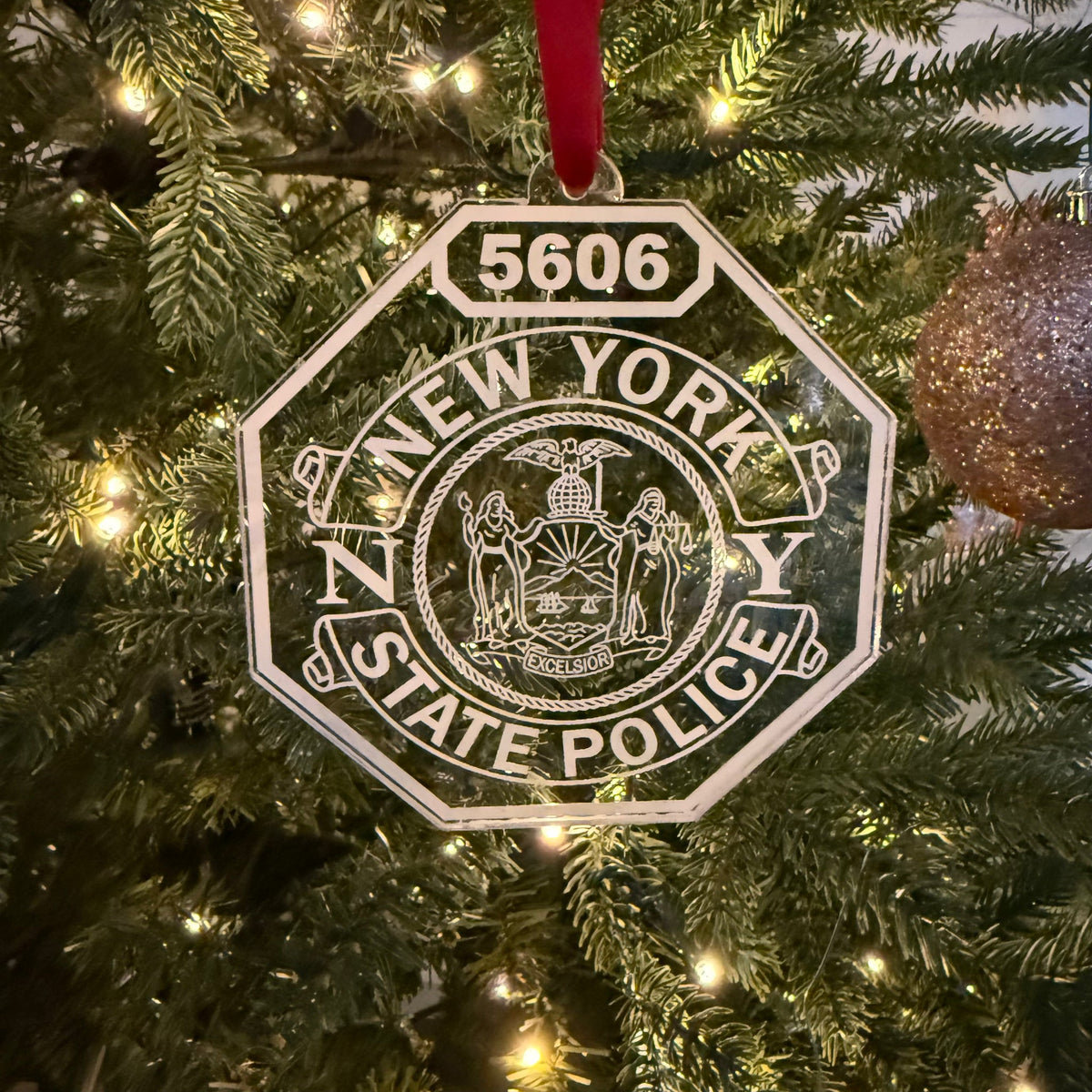 Clear acrylic New York State Police shield Christmas ornament engraved with shield number 5606, hanging from a red ribbon on a lit tree
